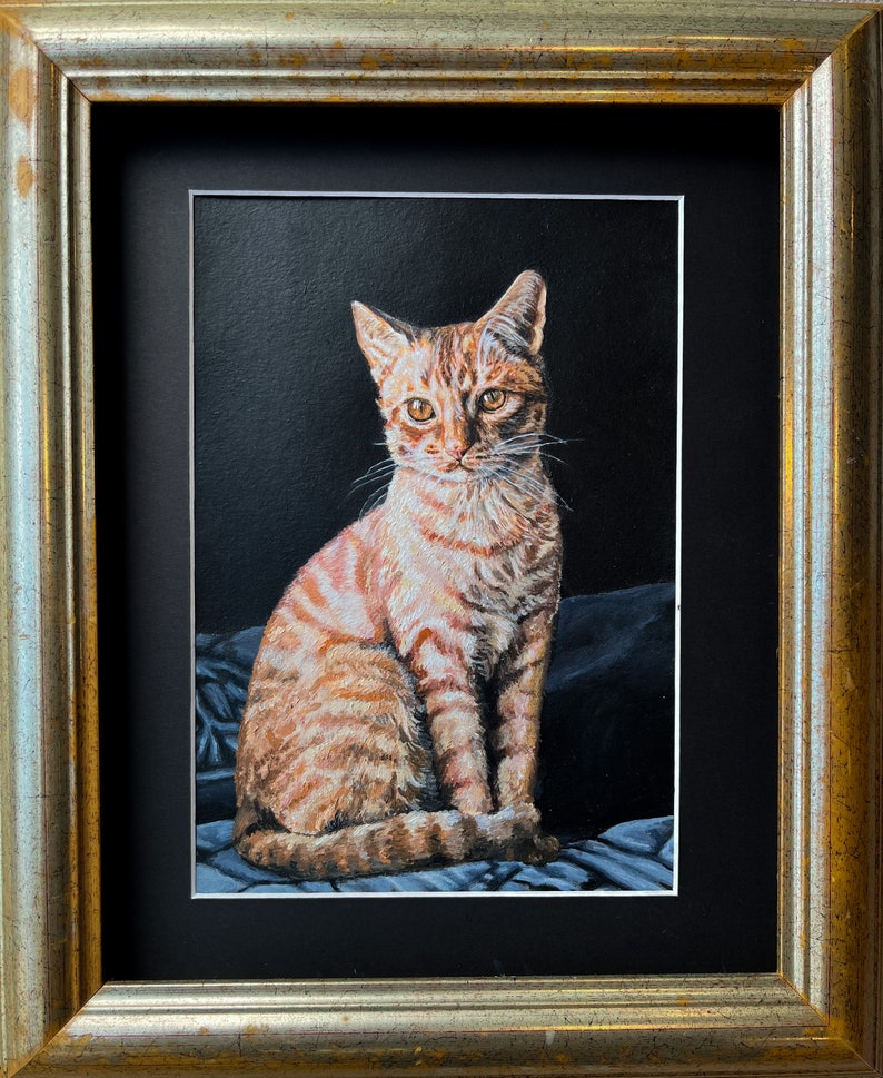 May include: A framed painting of a tabby cat sitting on a blue and white patterned fabric. The cat has orange and white fur and is looking directly at the viewer.