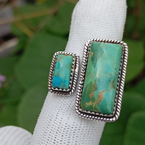 May include: Two silver rings with turquoise stones. The larger ring has a rectangular turquoise stone with a silver rope-like border. The smaller ring has a smaller rectangular turquoise stone with a similar border. The rings are displayed on a white surface.