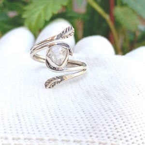 May include: A silver-toned ring with a clear, faceted gemstone at its center. The ring features a unique design with leaf-like accents on each side of the gemstone. The ring is displayed on a white surface, showcasing its intricate details.