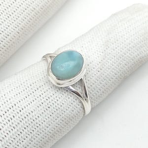 Könnte beinhalten: Ein Silberring mit einem hellblauen, ovalen Edelstein. Der Ring hat ein geteiltes Schaftdesign, wobei sich das Band auf beiden Seiten des Edelsteins in zwei separate Stränge aufteilt. Der Edelstein ist in einer Lünettenfassung gefasst.