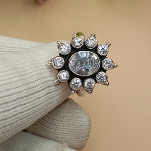 Op de afbeelding: Een zilverkleurige ring met een grote, ovaal geslepen heldere edelsteen in het midden, omringd door kleinere, ronde heldere edelstenen in een zonnestralenpatroon. De ring is geplaatst tegen een neutrale achtergrond.