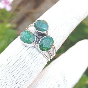 May include: A silver ring featuring three oval-cut emerald gemstones. The ring has a triple-band design and the emeralds are set in individual bezels. The gemstones are a vibrant green color.