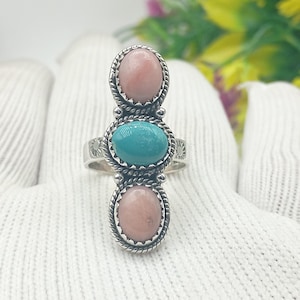 Op de afbeelding: Een zilveren ring met drie ovale edelstenen: twee roze en één turquoise. De edelstenen zijn gezet in een decoratieve, touwachtige rand. De ring heeft een gedetailleerde, gegraveerde band.