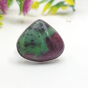 May include: A silver ring with a large, triangular gemstone. The gemstone has a mix of green and maroon colours, with a polished surface. The ring's band is simple, polished silver. The ring is set against a white background.