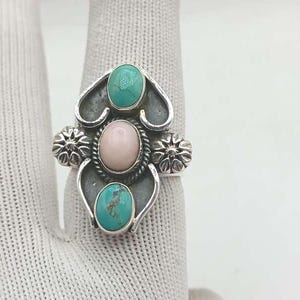 Puede incluir: Un anillo de plata con una piedra rosa ovalada central, flanqueada por dos piedras turquesas y detalles florales decorativos. El anillo presenta un diseño detallado y ornamentado, mostrando una mezcla de colores y texturas.