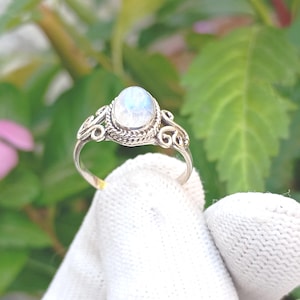 May include: A silver ring with an oval, iridescent moonstone. The ring features an ornate, filigree design around the stone and along the band. The ring is displayed against a backdrop of green leaves and a pink flower.