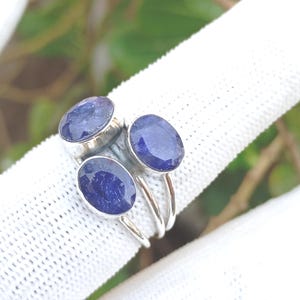 May include: A silver ring featuring three oval, deep blue gemstones. The gemstones are set in individual settings and arranged in a cluster design. The ring is displayed on a white fabric, with a blurred green background.
