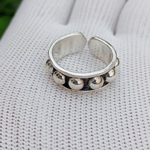 Puede incluir: Un anillo ajustable plateado con una fila de pequeñas cuentas redondas en relieve. El anillo tiene un diseño abierto, lo que permite ajustar la talla. El anillo se muestra sobre una superficie blanca.