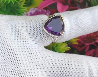 May include: A silver ring featuring a large, triangular, deep purple gemstone. The gemstone is set in a silver band, and the ring is displayed against a white, textured background with colorful flowers in the background. The ring is a piece of jewelry.