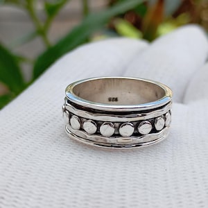 Puede incluir: Un anillo de plata con un diseño texturizado. El anillo presenta una banda de pequeños círculos espaciados uniformemente entre dos bandas texturizadas. El anillo está marcado con "925".
