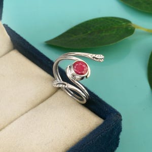 Puede incluir: Un anillo de plata con una piedra preciosa roja y redonda. El anillo tiene un diseño fluido en forma de hoja. El anillo se muestra en una caja de joyería azul oscuro, con un interior beige claro. El fondo es de color verde azulado claro con una hoja verde.