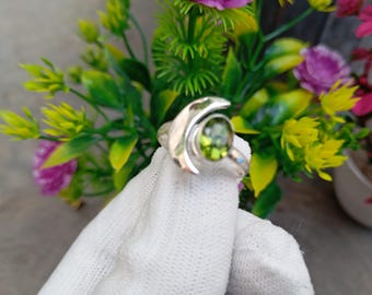 May include: A silver ring featuring a large, oval, light green gemstone. The ring has a unique design with a curved, polished band and a small, light blue accent stone. The ring is displayed against a backdrop of colorful flowers.