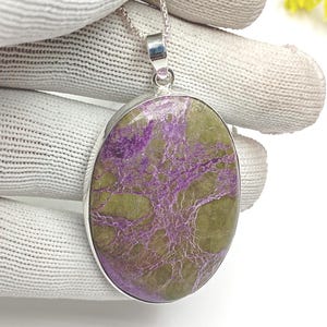 May include: Oval pendant necklace with a silver chain. The pendant features a large, polished stone with a mix of purple and olive green hues. The stone has a unique, natural pattern. The pendant is set in a silver bezel.