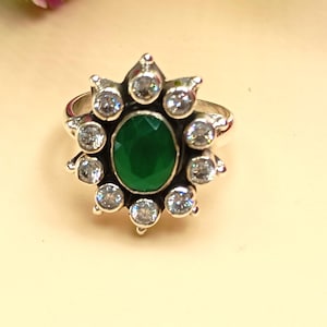 Op de afbeelding: Zilveren ring met een ovale, smaragdgroene steen in het midden, omringd door kleinere, heldere, ronde edelstenen. Het ontwerp van de ring doet denken aan een bloem of zonnestraal.
