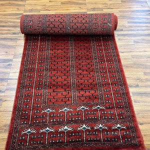 May include: A rolled-up, rectangular red rug with a detailed black and white pattern. The rug is partially unrolled, revealing a repeating design of vertical lines and stylized tree-like motifs. The rug has a white fringe on the end.