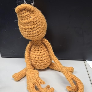 May include: A handmade, crocheted creature in shades of orange and black. The plush toy has a large head, a rounded body, and long, slender limbs. The eyes are black and white. The toy is sitting on a light gray surface.