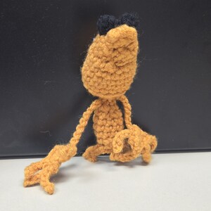 May include: A handmade crochet figure in shades of orange and black. The figure has a rounded head with two small black tufts, long arms, and a small body. The figure is posed with one arm extended and the other bent.