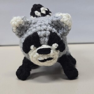 May include: A small, handcrafted crochet raccoon amigurumi featuring a grey body, black eye patches, a white muzzle, and black paws. The figure has pointed ears with white tips and a striped black and white tail extending from the back.