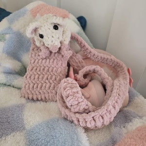 May include: A handmade crochet opossum toy in white and pink. The opossum has a pink scarf and a long, pink tail. It is lying on a soft, multi-colored blanket with pink, blue, and white squares. The toy is designed for children.