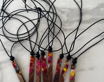 May include: A collection of handmade necklaces featuring rectangular wooden pendants inlaid with colorful resin. Each pendant is suspended from a black cord, with a black bead accent. The resin includes shades of red, pink, yellow, and turquoise.