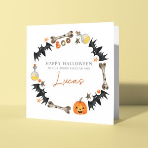 May include: A white Halloween card featuring a watercolour wreath design. The wreath includes bats, bones, stars, and a pumpkin. The text reads "Happy Halloween to our spooktacular son Lucas."