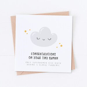 May include: A white card with a gray cloud with a smiling face and gold stars. The text on the card reads "Congratulations on your tiny human. Only superwomen give birth during a global pandemic."