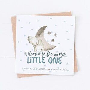 May include: A white card with a watercolor illustration of a gray elephant standing on a crescent moon with stars. The text "welcome to the world LITTLE ONE" is in blue. The card also includes the text "LOGAN PETER STEVENSON 30TH JUNE 2020".