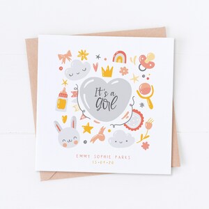 May include: A white card with a gray heart in the center that says "It's a girl". The heart is surrounded by a variety of colorful illustrations, including a crown, a pacifier, a baby bottle, a cloud, a bunny, and a star. The card also includes the text "EMMY SOPHIE PARKS 13-04-20".