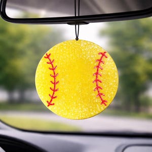 May include: A yellow softball-shaped car air freshener with red stitching detail, hanging from a black string. The air freshener is suspended from a car's rearview mirror, with a blurred outdoor background.