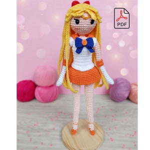 Pode incluir: Boneca de crochê com cabelo amarelo, saia laranja, top branco e laço azul. A boneca está em pé sobre uma base de madeira.