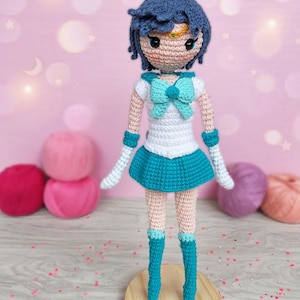 Pode incluir: Boneca Ami Mizuno de crochê de Sailor Moon. A boneca tem uma peruca azul, uma parte superior branca com um laço azul e uma saia azul. A boneca está em uma base de madeira. O texto "Ami Mizuno - Sailor Mercury" está no topo.