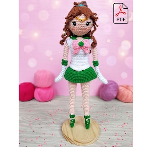 Puede incluir: Muñeca de crochet con cabello castaño, falda verde, camisa blanca y lazo rosa. La muñeca está de pie sobre una base de madera.