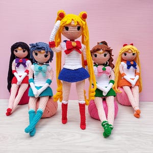 Pode incluir: Cinco bonecas de crochê feitas à mão, cada uma com roupas e penteados únicos. As bonecas apresentam cores vibrantes como azul, vermelho, amarelo, verde e laranja. Elas estão sentadas em bolas de fio rosa, mostrando um design caprichoso e artístico.