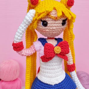 Pode incluir: Boneca de crochê feita à mão de uma personagem com cabelo amarelo estilizado em dois coques com detalhes vermelhos. A boneca usa uma roupa de marinheiro branca, azul e vermelha com um grande laço vermelho. A boneca está em um fundo rosa.