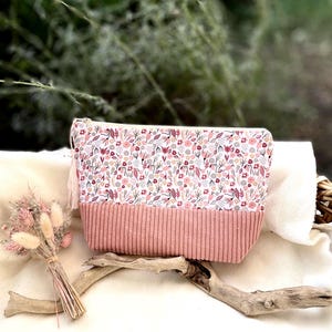 Può includere: Una pochette con cerniera caratterizzata da una sezione superiore su sfondo bianco con un motivo floreale multicolore nei toni del rosa, rosso e grigio. La sezione inferiore è realizzata in velluto a coste rosa polvere con trama verticale.