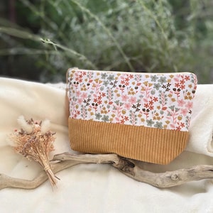 Peut inclure: Une pochette zippée rectangulaire avec une partie supérieure blanche ornée d'un motif floral multicolore dans des tons de rose, vert et marron. La partie inférieure est en tissu velours côtelé jaune moutarde avec une texture striée verticale.