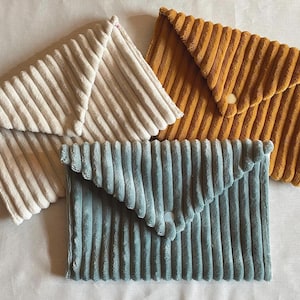 Può includere: Tre pochette a busta in velluto a coste color crema, giallo senape e verde acqua. Le pochette hanno una texture a coste e una chiusura a patta triangolare. La pochette giallo senape ha una chiusura con bottone. Le pochette sono disposte su una superficie bianca.