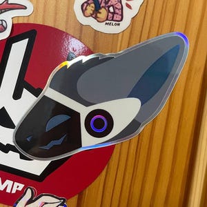 Puede incluir: Una pegatina con la imagen de una cabeza de protógeno estilizada en negro, gris y blanco. La pegatina tiene un efecto holográfico y está colocada sobre una superficie de madera con otras pegatinas.