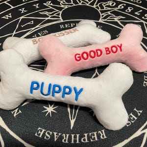 Könnte beinhalten: Drei Plüsch-Hundespielzeug in Knochenform mit besticktem Text. Der weiße Knochen sagt "Puppy" in Blau, der rosa Knochen sagt "Good Boy" in Rot und der weiße Knochen sagt "Boy Kisser" in Gold.
