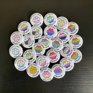 Op de afbeelding: Een verzameling van 20 ronde witte knoppen met poot afdrukken en tekst. Elke knop heeft een andere LGBTQ+ identiteitsvlag en de woorden "en trots".