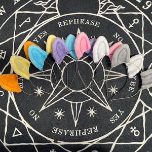 May include: A set of nine colourful fuzzy bunny ear headbands. The headbands are arranged in a circle on a black background with a white star pattern. The headbands are in the following colours: orange, yellow, light blue, purple, pink, white, grey, light grey, and dark grey.