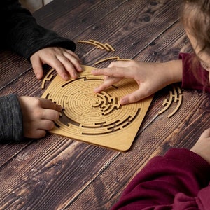 May include: Two people working together to solve a wooden maze puzzle. The puzzle is made of brown wood and has a circular design. The maze is intricate and challenging.