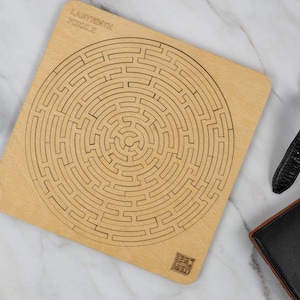 May include: A wooden labyrinth puzzle with a circular design. The puzzle is made of light brown wood and has a natural finish. The text "Labyrinth Puzzle" is printed on the top of the puzzle.