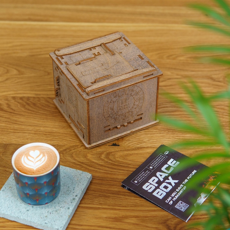 May include: A wooden box with the words "Space Box" on the front. The box is sitting on a wooden table with a cup of coffee and a pamphlet that reads "Can you save the future of humanity?"