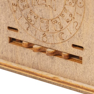 May include: Close-up of a wooden box with a circular design and a gear mechanism. The design features symbols and numbers, possibly representing a calendar or a clock.
