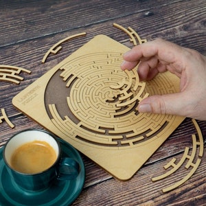 May include: A wooden laser cut labyrinth puzzle with the words "Labyrinth Puzzle" printed on the top. A hand is holding a piece of the puzzle, which is a spiral shape.