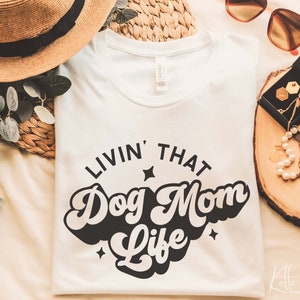 May include: White t-shirt with black text that reads "Livin' That Dog Mom Life" in a retro font. The text is surrounded by small stars.
