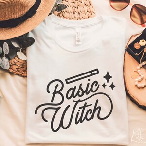 May include: White t-shirt with the text "Basic Witch" in a black cursive font. A small black wand is above the text with two small black stars on either side.