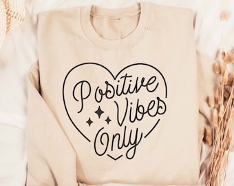 Peut inclure: Un sweat-shirt beige avec un motif de cœur noir qui dit "Positive Vibes Only" avec trois petites étoiles à l'intérieur du cœur.