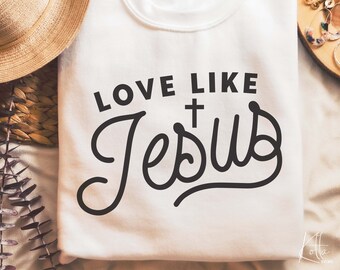 Puede incluir: Sudadera blanca con el texto "Love Like Jesus" en letras negras con un símbolo de cruz sobre la palabra "Jesus".